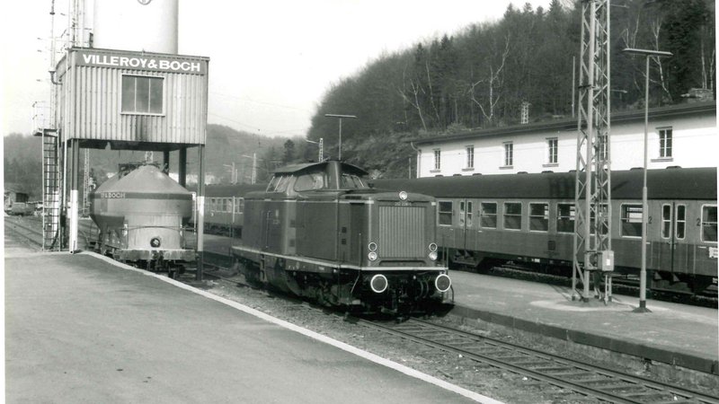 schwarz-weiß Fotografie von 1971. Es ist eine Lok im Bahnhofsbereich zu sehen. Auf den Schienen dahinter ist ebenfalls ein Zug mit mehreren Waggons zu sehen.