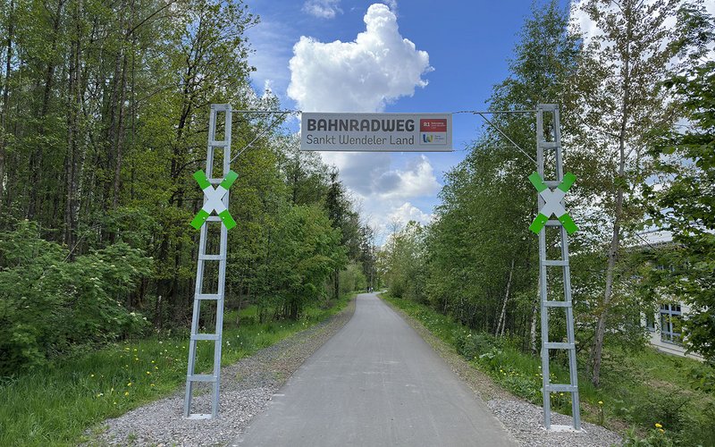 Das Eingangstor am Bahnradweg besteht aus 2 hohen Eisengestängepfosten, die mit einem Drahtseil verbunden sind. An diesem Drahtseil ist ein Banner befestigt mit der Aufschrift Bahnradweg Sankt Wendeler Land und dem Logo des Weges. An den beiden Eisenpfosten ist jeweils ein grün-weißes Andreaskreuz aufgehängt..  