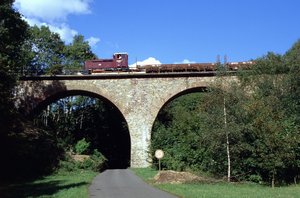 über das Nonnweiler Viadukt fährt die Museumsbahn mit ihrem roten Zug und mehreren Waggons. An den Bildrändern sind Bäume und Büsche zu sehen. Der Himmel ist kaum bewölkt und klar. 