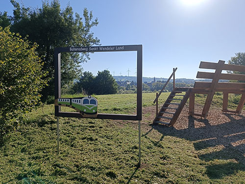 Landschaftsaufnahme mit einem großen Holzfotorahmen mit der Aufschrift Bahnradweg Sankt Wendeler Land. Neben diesem Fotorahmen ist eine überdimensional große XXL-Bank zu sehen. Ein Leiteraufgang führt zur Sitzfläche der Bank von der man einen Blick über die Landschaft genießen kann.